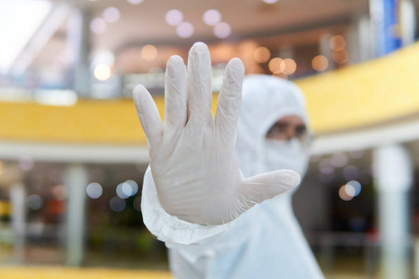 человек в покрывающем одноразовом противоэпидемическом антибактериальном изоляционном костюме демонстрирует категорический жест запрета на фоне некоторого интерьера общественного пространства
 - Фото, изображение