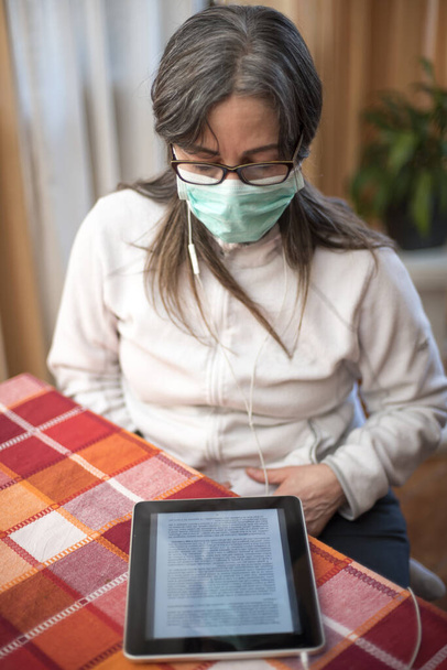 Het Corona Virus. Zieke vrouw van het corona-virus die op de tablet leest, die ademhalingsbescherming draagt en herstelt van de ziekte thuis. Patiënt geïsoleerd om infectie te voorkomen - Foto, afbeelding
