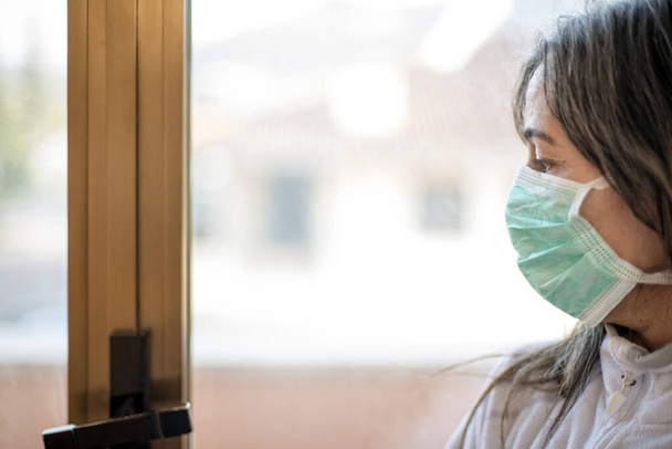 Corona Virus. Nemocná žena s virem korony se dívá oknem a má na sobě ochranu dýchacích cest a zotavuje se z nemoci doma. Pacient izolován, aby se zabránilo infekci - Fotografie, Obrázek