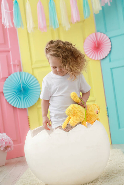Ένα μικρό παιδί μικρό παιδί κάθεται σε ένα λευκό αυγό που εκκολάφθηκε σε ένα στούντιο πολύχρωμο φόντο για ένα νεογέννητο πορτρέτο ή οικογενειακή ιδέα. - Φωτογραφία, εικόνα
