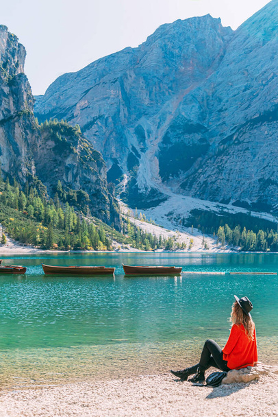 Happy tourist woman in red sweater brown long hair black hat sits on shore of Italy lake Braies. Turned around resting enjoys boats on turquoise water mountains covered forest backdrop divine nature - Photo, Image