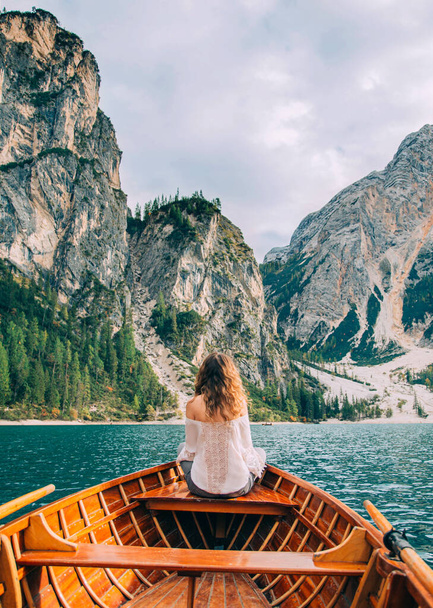 Redhaired girl turned away, sitting in the boat. Tourist in white blouse bare shoulders enjoy beauty alpine lake Braies backdrop magical nature dolomite mountains covered green forest. Vertical image - Foto, Imagen