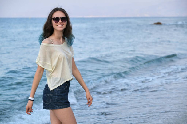 Trendy beautiful teenager girl with long colored blue hair in sunglasses and shorts walking along sea beach. Sea sunset, copy space - Fotografie, Obrázek