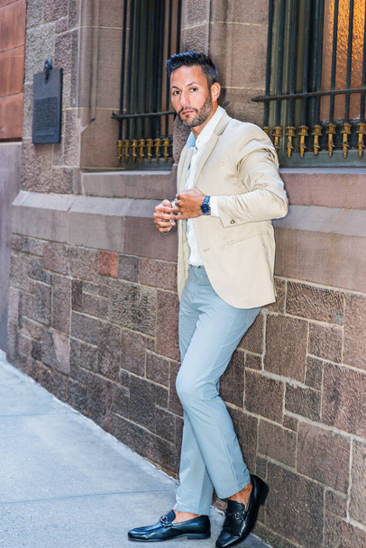 Jonge Europese zakenman met baard ontspant zich buiten in New York City, draagt beige blazer, grijze broek, zwarte leren schoenen, staat tegen een muur met ramen op straat, kijkt weg, denkt na - Foto, afbeelding