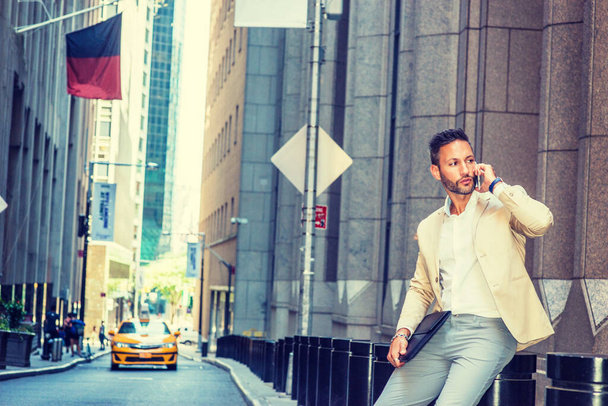 Joven Empresario Europeo viajando, trabajando en la ciudad de Nueva York, con barba, poco pelo gris, vistiendo chaqueta beige, pantalones grises, portando maletín, sentado en la calle vieja, hablando por teléfono celular
 - Foto, Imagen
