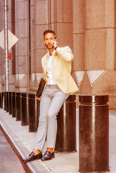Empresario europeo viajando, trabajando en la ciudad de Nueva York, con barba, poco pelo gris, vistiendo chaqueta beige, pantalones grises, zapatos de cuero, portafolios, sentado en la calle, hablando por teléfono celular
 - Foto, Imagen