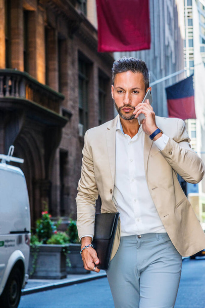 Young European Businessman traveling, working in New York City, with beard, little gray hair, wearing beige blazer, holding briefcase, walking on street, talking on cell phone, looking down, thinking - Photo, Image