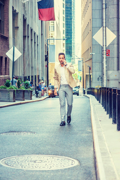 Jonge Europese zakenman reizend, werkend in New York City, met baard, klein grijs haar, beige blazer dragend, grijze broek, zwarte leren schoenen, lopend op smalle straat, pratend op mobiele telefoon - Foto, afbeelding