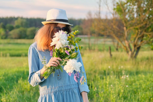 Beautiful teenager girl in jeans dress hat with bouquet of wildflowers, background scenic landscape, beauty, nature, copy space - Foto, afbeelding