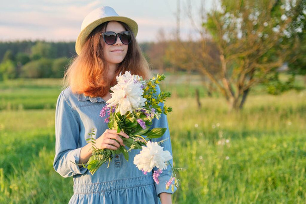 Portrait of young smiling beautiful girl in sunglasses hat with bouquet of wildflowers, sunset in meadow, copy space, rural scenery, golden hour - Foto, immagini