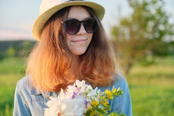 Portrait of young smiling beautiful girl in sunglasses hat with bouquet of wildflowers, sunset in meadow, copy space, rural scenery, golden hour - Foto, imagen