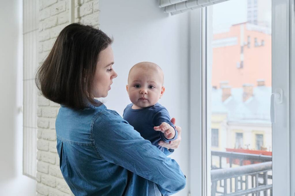Portrait of young beautiful mother and toddler son, woman holding baby in arms standing at home near window, copy space - 写真・画像