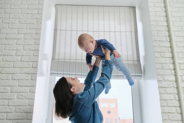 Jovem mãe bonita feliz brincando com o filho da criança, mulher levantando o bebê, voando e rindo menino. A família está feliz em casa, perto da janela, espaço de cópia
 - Foto, Imagem