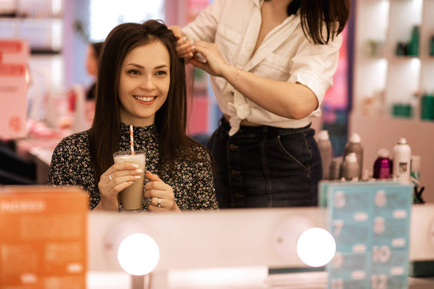 žena kadeřnice dělá účes k brunetce v salonu krásy, proces vytváření účes, profesionální kadeřník vytváří objem vlasů a klient dívka se usmívá a pije kávu - Fotografie, Obrázek