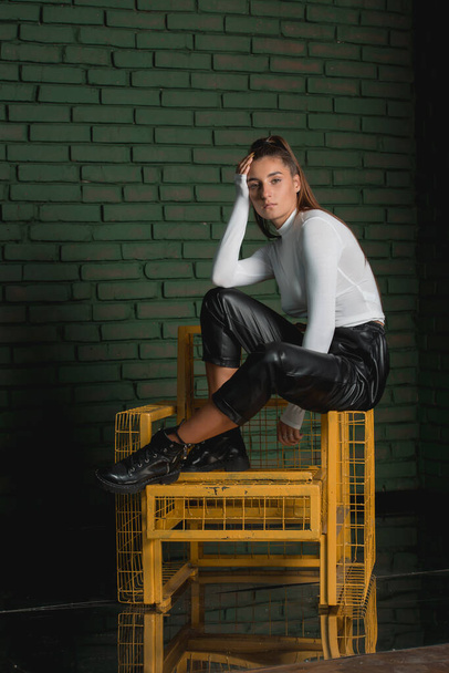 Fashionable brunette girl in a white sweater and black leather pants posing at the sunshine on an green bricks background. - Фото, изображение