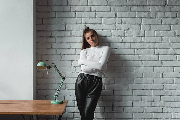 Fashionable brunette girl in a white sweater and black leather pants posing at the sunshine on an white bricks background. - Photo, Image