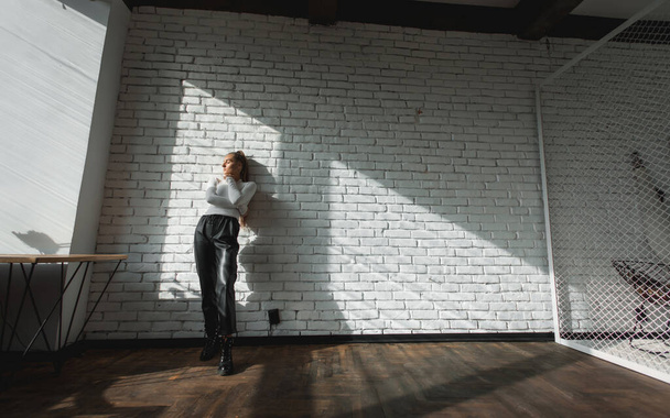 ファッショナブルなブルネット女の子でAホワイトセーターと黒革パンツで太陽の光でザ · ホワイトレンガの背景. - 写真・画像