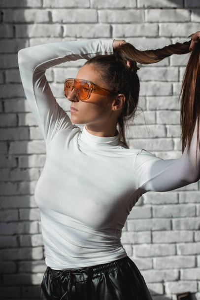 ファッショナブルなブルネットの女の子でAホワイトセーター,オレンジ色の眼鏡と黒革のパンツポージング上の白いレンガの背景. - 写真・画像