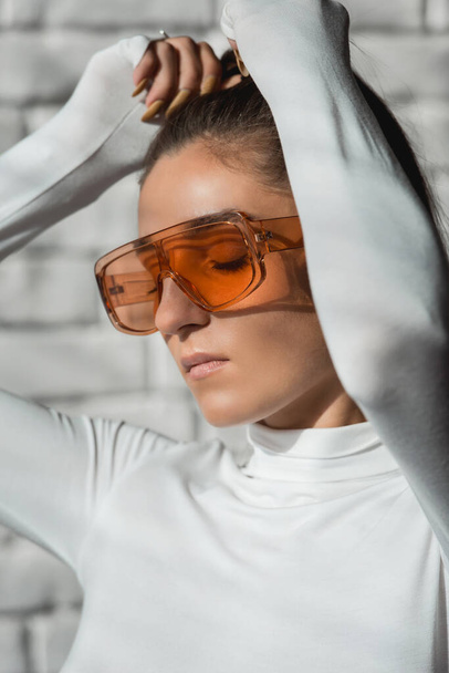 Fashionable brunette girl in a white sweater, orange glasses and black leather pants posing on an white bricks background. - Fotografie, Obrázek
