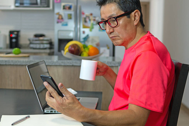 Wer zu Hause arbeitet, checkt sein Handy, während er mit seinem Computer in der Küche Kaffee trinkt - Foto, Bild