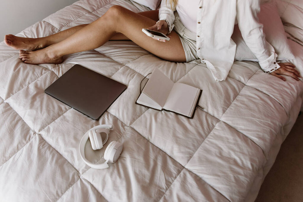 白人ラテン系白人の少女が携帯電話を使ってベッドに座ってた。フリーランスは自宅で働いています。ベッドの上に携帯電話ノートパソコンノートパソコンノートパッドヘッドフォン. - 写真・画像