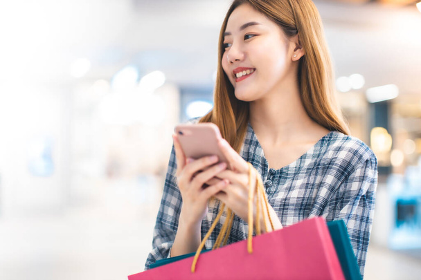 Χαμογελώντας νεαρή Ασιάτισσα γυναίκα με τσάντες χρώμα ψώνια πάνω από το εμπορικό κέντρο φόντο. χρησιμοποιώντας ένα έξυπνο τηλέφωνο ψώνια σε απευθείας σύνδεση και χαμογελώντας, ενώ στέκεται κτίριο εμπορικό κέντρο. έννοια τρόπου ζωής - Φωτογραφία, εικόνα