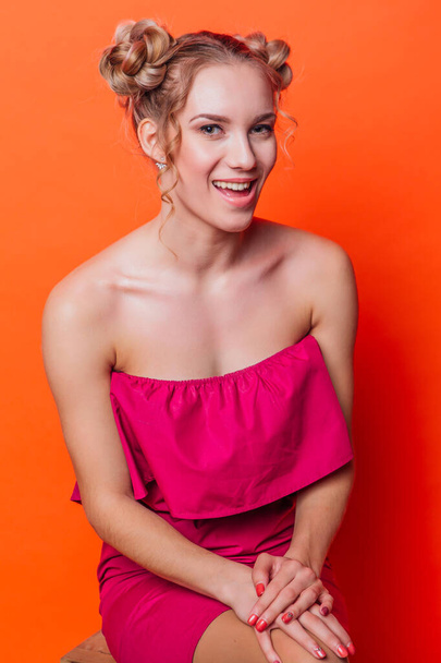 The girl in a red dress on a orange background in the studio. Blonde girl with two hair knots holding hands on knees, looking to the camera and smiling. - Photo, Image