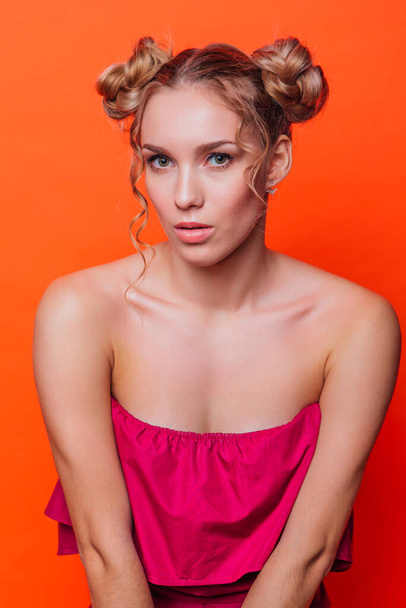 The girl in a red dress on a orange background in the studio. Blonde girl with two hair knots holding hands on knees, looking to the camera and smiling. - Foto, imagen