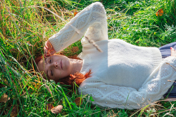 Morning portrait of a beautiful happy red woman with red hair laying in grass in white sweater - Zdjęcie, obraz