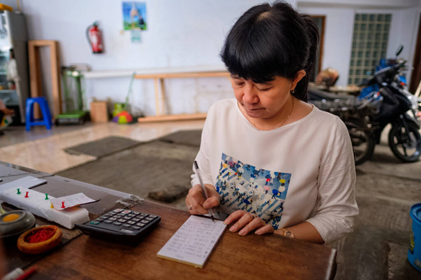 Ασιάτισσες Ινδονήσιες, ιδιοκτήτρια ενός μικρού τοπικού οικογενειακού καταστήματος, ή τοπικά αποκαλούμενου warung, υπολογίζοντας το κέρδος από το γραφείο της. Επιλογή εστίασης. - Φωτογραφία, εικόνα