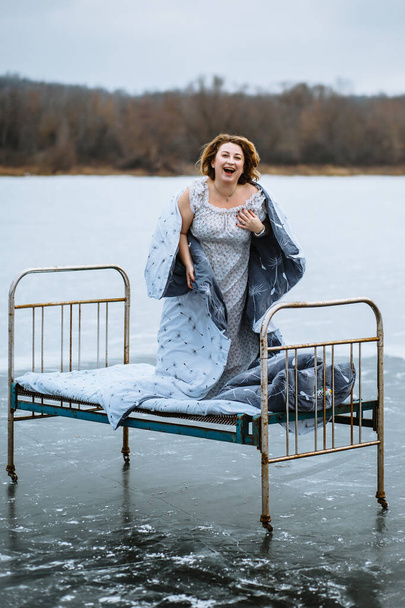 A girl in a nightgown and a blanket , waking up on a bed on a frozen blue lake - Фото, изображение