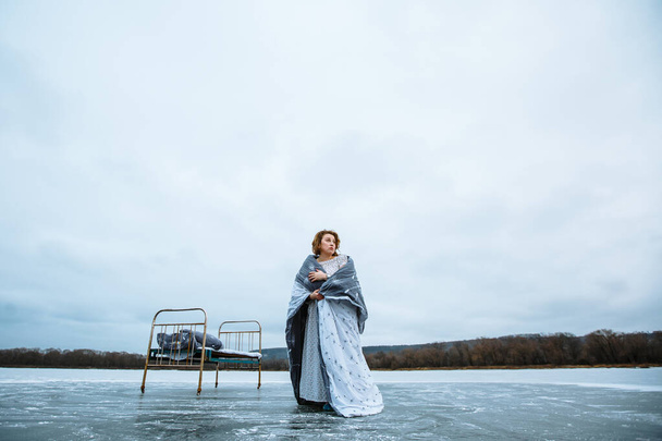 A girl in a nightgown and a blanket standing on the ice of a blue lake, next to an iron bed - Fotografie, Obrázek