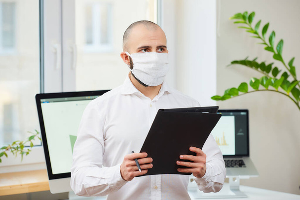 Orvosi maszkot viselő férfi a koronavírus ellen (COVID-19). Egy mérnök tollal és egy mappával a munkahelyén, számítógépekkel és zöld növényekkel a háttérben. Koronavírus-karantén. - Fotó, kép
