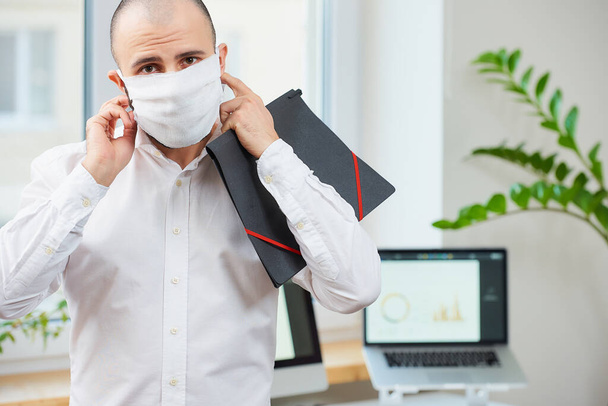 Koronavirüse karşı tıbbi prosedür maskesi çıkaran bir adam. Çalışma alanında elinde bilgisayarları ve arka planda yeşil bitkileri olan bir dosya tutan bir ofis çalışanı. COVID-19 karantinası. - Fotoğraf, Görsel