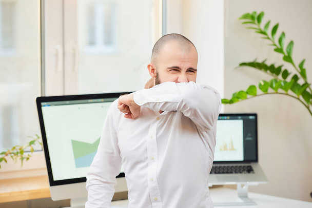 Un homme en chemise blanche avec une barbe tousse fortement dans le coude. Un employé de bureau à son espace de travail avec des ordinateurs et des plantes vertes en arrière-plan. Quarantaine de coronavirus (COVID-19)
. - Photo, image
