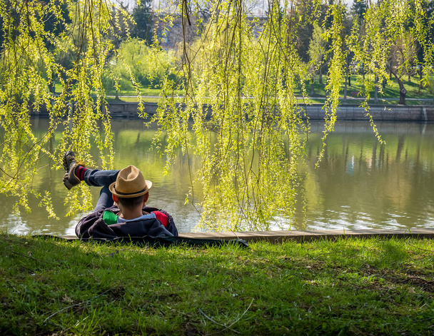 Een man met een hoed op het gras aan de rand van een meer. Een man die geniet van een mooie dag in het park. - Foto, afbeelding