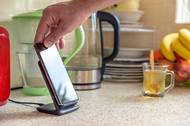 smartphone wireless charging on charging stand on kitchen tabletop. male hand placing phone to charging - Foto, afbeelding