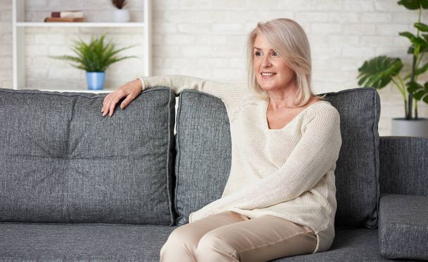 Portrait of senior woman is sitting on a sofa in living room - Фото, изображение