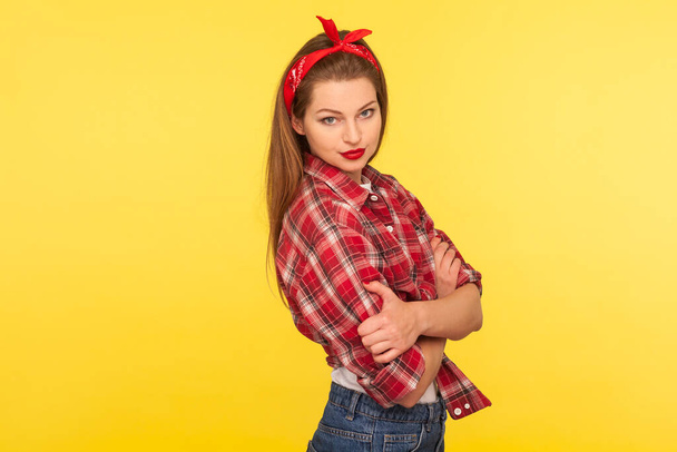 Portrait of sexy pinup girl with bright makeup, red lipstick, wearing checkered shirt and headband, standing with crossed hands and looking seductively, retro vintage 50's style. studio shot isolated - Photo, Image