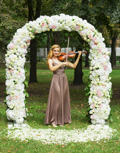 Молода дівчина в довгій сукні грає на скрипці. усміхнена дівчина в весільній арці
. - Фото, зображення