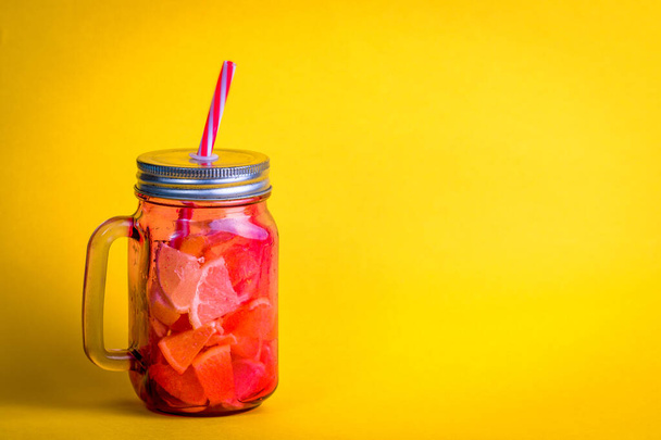 Rode glazen kruik met handvat met koude natuurlijke drank met stukjes fruit en gele achtergrond. - Foto, afbeelding