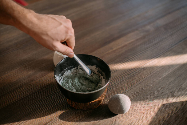 Face and body care. Preparation of a cosmetic mask in a bowl on a wooden table. Men's hands make a face mask from powder and water. - Zdjęcie, obraz