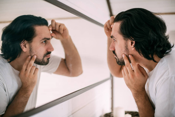 Un hombre guapo se mira en el espejo. El cuidado personal de los hombres. Un joven se examina a sí mismo en el espejo del dormitorio. - Foto, imagen