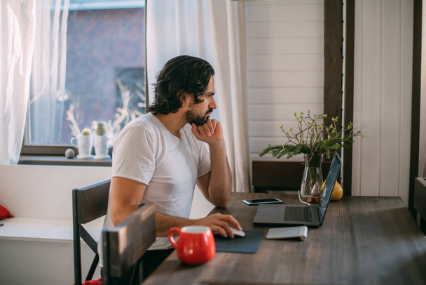 Munkahely otthon. Egy férfi otthon dolgozik egy laptopnál, napközben egy asztalnál ül a nappaliban. Otthoni munka. - Fotó, kép