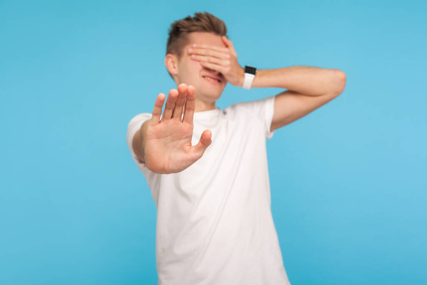 I'm not watching! Portrait of scared shamed man in casual white t-shirt covering eyes and showing stop gesture, hiding eyesight, feeling shy to watch. indoor studio shot isolated on blue background - Foto, Imagen