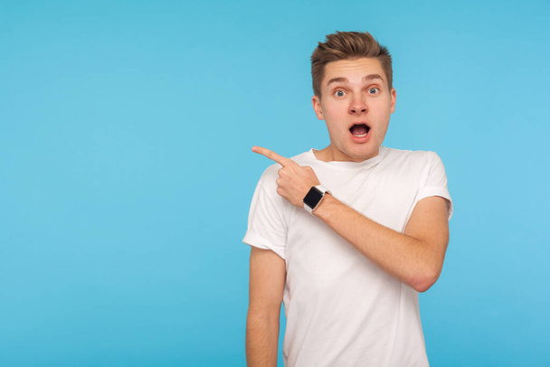 Wow look, advertise here! Portrait of young man expressing amazement and shock while pointing to the side, showing blank copy space for crazy idea presentation. studio shot isolated on blue background - 写真・画像