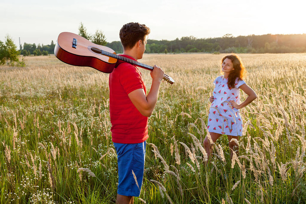 A young man with the guitar is standing near his pregnant wife in the wheat field on a sunny day.Pregnant family photo shoot in nature - Foto, Imagem