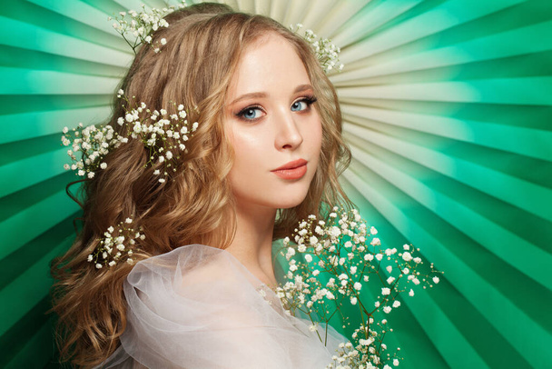 Cute young woman with long healthy blonde curly hair, make up and flowers on green background - Zdjęcie, obraz
