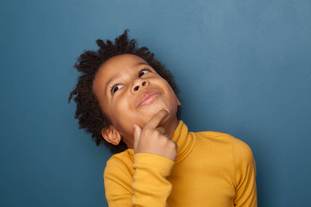 Niño negro pequeño pensando y mirando hacia arriba sobre fondo azul
 - Foto, Imagen