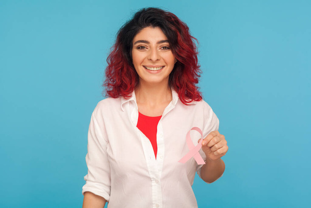 Weibliche Gesundheit. Positive Frau mit ausgefallenen roten Haaren, die in die Kamera lächelt und eine rosafarbene Schleife in der Hand hält, Symbol des Brustkrebsbewusstseins, der Diagnostik und Prävention onkologischer Erkrankungen. Innenaufnahmen aus dem Studio - Foto, Bild